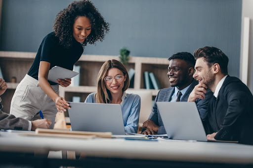 A diverse group of business professionals collaborates around a laptop, discussing AI implementation and practical solutions. This image represents the collaborative and business-focused nature of SADA Ground School's approach to technology.
