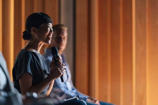 A woman speaking confidently into a microphone at a conference, representing thought leadership and expert guidance. This image signifies the authority and expertise of SADA's presenters, who provide the tools and knowledge to navigate the challenges of AI adoption